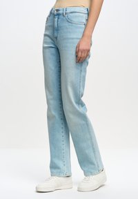 Jeans de mezclilla azul claro con un diseño de pierna recta, que cuentan con un cierre de botón y cremallera clásico, bolsillos laterales y detalles de costura. Combinados con zapatillas blancas.