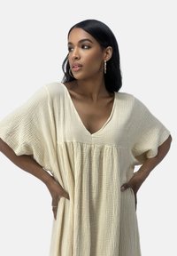 Beige jurk met structuur, diepe V-hals, korte mouwen en getailleerde plooien bij de taille, met een losse, vloeiende pasvorm.