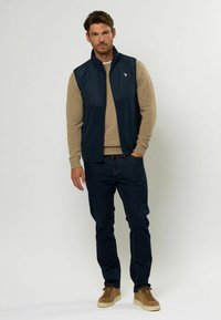 Marinblå väst med dragkedja fram, kombinerad med en beige tröja och mörka jeans. Skorna är bruna mockasneakers. Enkel, funktionell design.