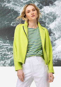 Neon groene, zip-up jas met een getailleerd ontwerp. Gecombineerd met een blauw-groene gestreepte shirt. Model draagt witte broeken. Abstracte oceaan achtergrond.