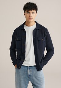 Marinblå stickad cardigan med knäppning framtill, två bröstfickor, rundad krage och ribbade ärmslut, bärs över en vit T-shirt.