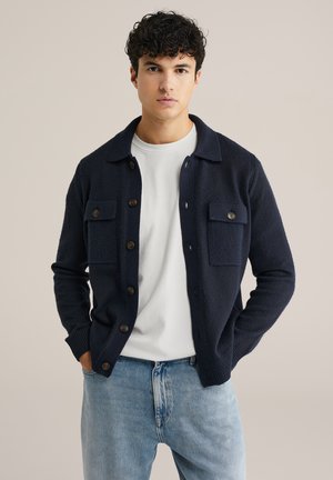 Marinblå stickad cardigan med knäppning framtill, två bröstfickor, rundad krage och ribbade ärmslut, bärs över en vit T-shirt.