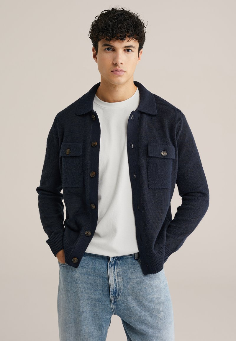 Marinblå stickad cardigan med knäppning framtill, två bröstfickor, rundad krage och ribbade ärmslut, bärs över en vit T-shirt.