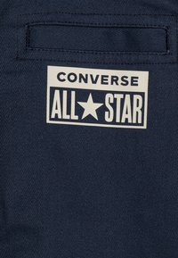 Granatowa płótno z teksturowanym wykończeniem, z białą, prostokątną etykietą z napisem "CONVERSE ALL★STAR" oraz ikoną gwiazdy.
