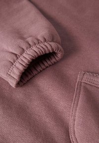 Tessuto di felpa mauve chiaro con un motivo a maglia testurizzata, caratterizzato da un polsino arricciato e una piccola tasca sul davanti. Texture liscia ed elastica.