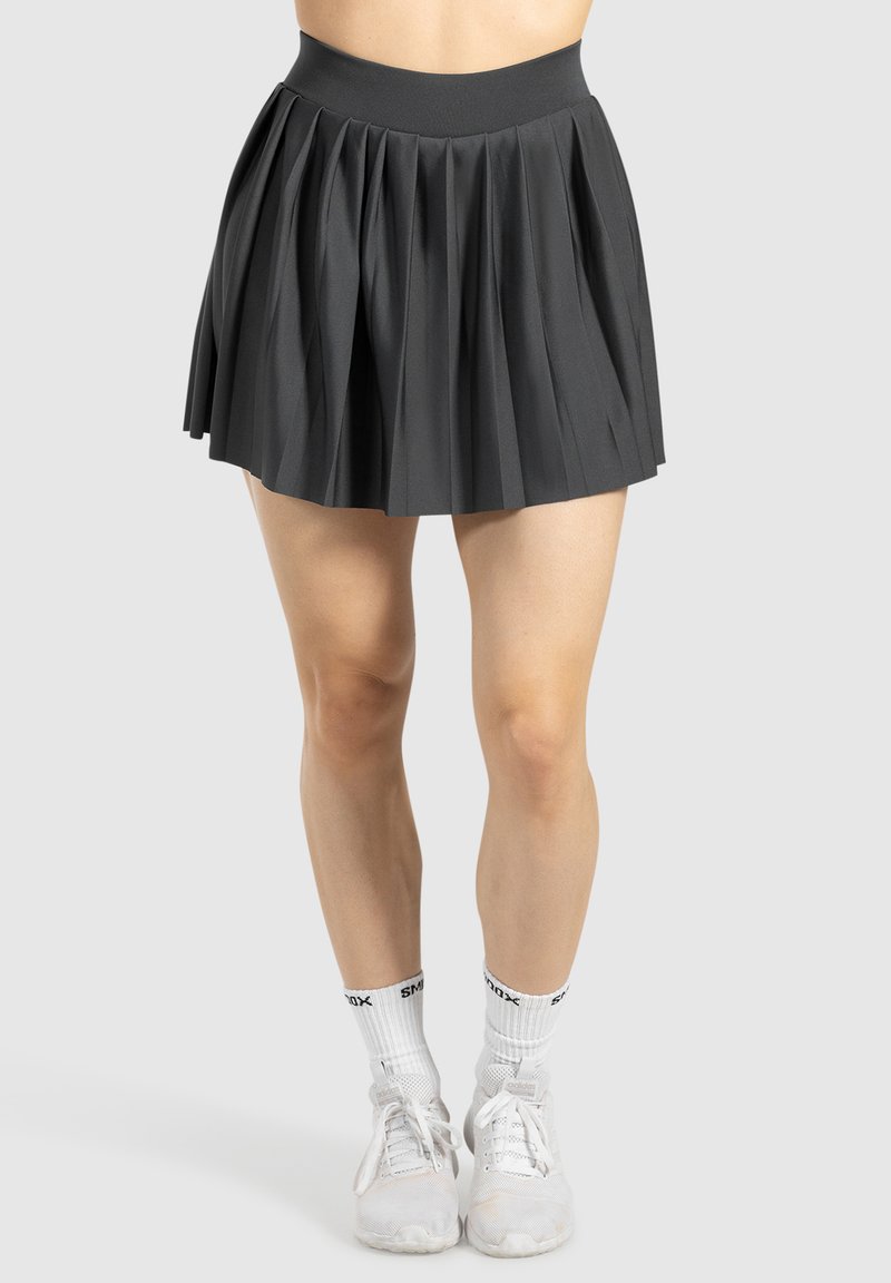 Zwarte geplooide rok met een hoge tailleband, gemaakt van een textuurstof. Witte sportschoenen en sokken maken de outfit compleet. Eenvoudig, modern ontwerp.