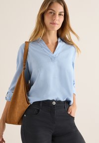 Blusa azzurro chiaro a maniche lunghe con scollo a V e polsini arrotolati, abbinata a pantaloni blu scuro. È anche visibile una borsa a spalla in camoscio marrone.