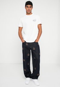 Hvit bomulls T-skjorte med et logo på brystet, kombinert med mørkeblå jeans som har fargerike broderte krone-design og hvite joggesko.