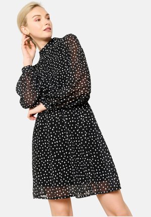 Robe noire à pois blancs en tissu transparent, avec un col haut, des manches longues bouffantes et une taille élastique. Présente une longueur fluide, au-dessus du genou.