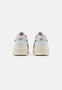 Zapatillas gris y blancas con acentos rosas y una marca plateada texturizada. Cuenta con un collar acolchado y una suela de goma acanalada. Líneas de diseño limpias.