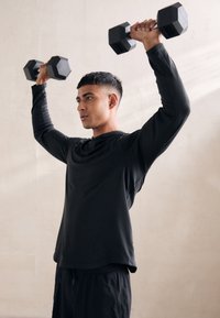 Schwarzes Langarm-Sportshirt und Shorts, getragen von einer Person, die schwarze sechseckige Kurzhanteln über dem Kopf hebt, vor einem hellen Hintergrund.