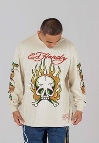 Beige Langarmhemd mit Totenkopf- und Flammengrafik; bunte Tattoos auf den Ärmeln; Text "Ed Hardy" in Rot über dem Design.