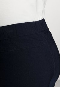 Detailní záběr na osobu oblečenou v tmavě modrých elastických kalhotách s páskem a bílém tričku zastrčeném do kalhot na jednobarevném pozadí.