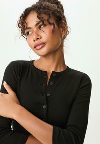 Schwarzes langärmliges Henley-Oberteil mit rundem Ausschnitt, Knopfdetails und weichem Material, präsentiert an einer Person mit lockigem Haar.