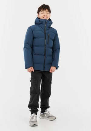 Veste matelassée bleu marine avec col montant, fermeture éclair à l'avant et poche poitrine, associée à un pantalon cargo noir et des baskets grises.