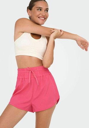 Lyserøde atletiske shorts med elastisk talje og snor. En ærmeløs cremefarvet crop top bæres over shortsene.