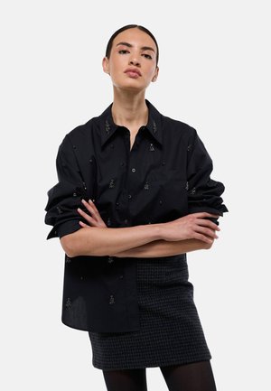 Chemise noire avec un design à boutons, des accents brodés et des manches roulées. Portée par-dessus une jupe à carreaux sombre.