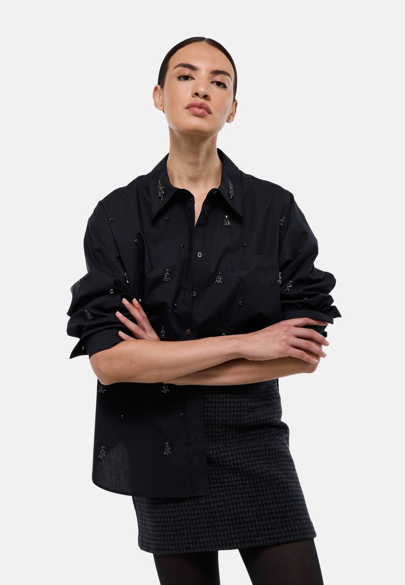 Chemise noire avec un design à boutons, des accents brodés et des manches roulées. Portée par-dessus une jupe à carreaux sombre.