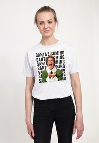 Bílá bavlněná tričko s grafickým potiskem muže s kudrnatými vlasy, oblečeného v zeleném oblečení s bílými akcenty, a textem "SANTA PŘICHÁZÍ."