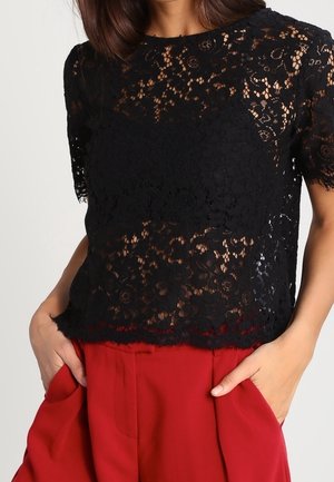 Vrouw draagt een zwarte korte top met bloemenkant en ronde hals, gecombineerd met hoge rode broek met handen in de zakken.