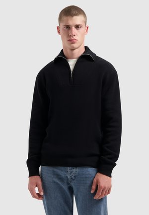 Jonge man in een zwart half-rits trui over een wit shirt en blauwe jeans, staande tegen een effen lichtgrijze achtergrond.