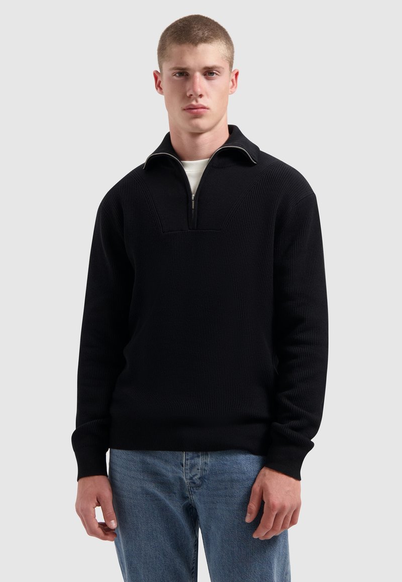 Jonge man in een zwart half-rits trui over een wit shirt en blauwe jeans, staande tegen een effen lichtgrijze achtergrond.