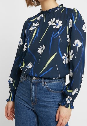 Blouse bleue marine à imprimé floral blanc, col montant à volants et poignets, portée rentrée dans un jean bleu taille haute.