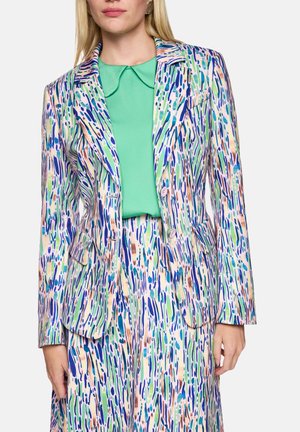 Woman wearing colorful abstract patterned suit jacket and matching skirt with a green blouse underneath, standing with hands at sides.