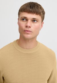 Beige gerippter Pullover mit rundem Ausschnitt und strukturiertem Strickmuster. Das Model hat kurze braune Haare und blaue Augen. Einfarbiger Hintergrund.