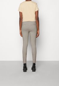 Personne portant une chemise beige à manches courtes, un pantalon slim à carreaux et des bottines à talons noires, debout devant un arrière-plan blanc uni.