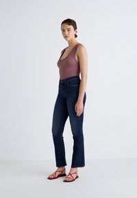 Een aansluitend geribbeld tanktop in mauve, gecombineerd met donkerblauwe high-waisted flared jeans en rode bandjes sandalen. Eenvoudige achtergrond.