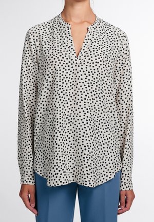 Donna che indossa una camicetta bianca a maniche lunghe con pois neri irregolari abbinata a pantaloni blu a vita alta, in piedi su sfondo bianco.