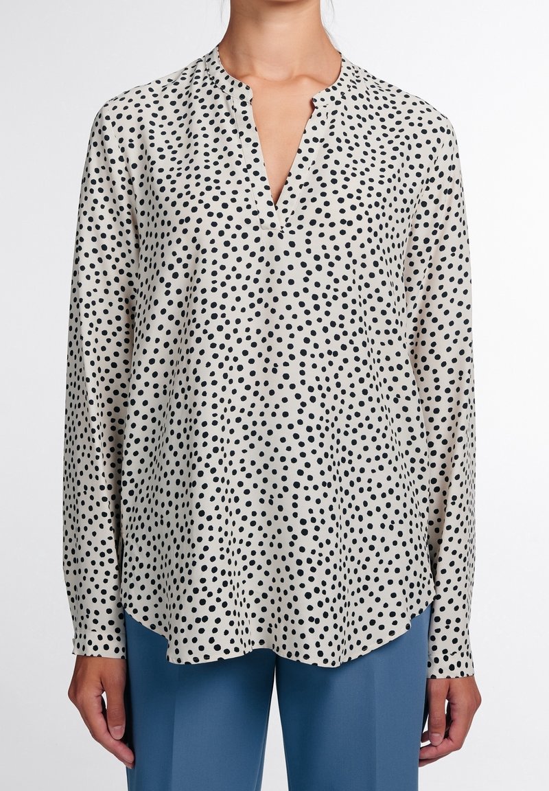 Femme portant un chemisier blanc à manches longues avec des pois noirs irréguliers, associé à un pantalon bleu taille haute, debout sur fond blanc.