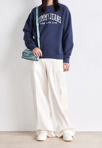 Tummansininen huppari, jossa on teksti "TOMMY JEANS NEW YORK CITY", valkoiset löysät cargo-housut taskuilla ja valkoiset lenkkarit; pitämässä teal-väristä olkalaukkua.