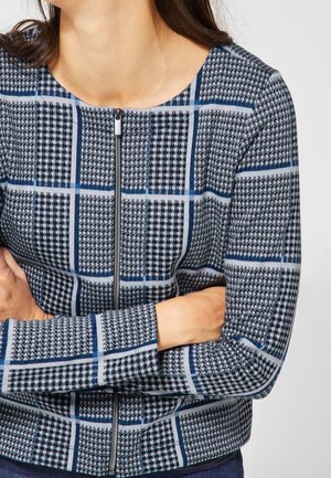 Kobieta w niebiesko-czarnej, kraciastej kurtce zapinanej na zamek, z długimi rękawami, ze skrzyżowanymi ramionami, na jednolitym tle.