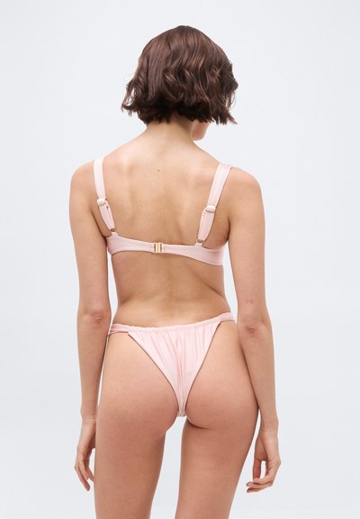 Γυναίκα με κοντά καστανά μαλλιά που φοράει ένα ανοιχτό ροζ μπικίνι, εμφανιζόμενη από πίσω σε απλό λευκό φόντο.