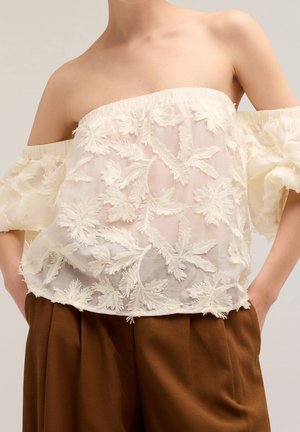 Haut blanc à col bardot avec broderie florale, texture transparente et manches à volants, associé à un pantalon marron taille haute.