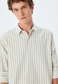Camisa de botones a rayas en tonos beige claro y gris, con cuello, mangas cortas y un corte holgado con una textura de tela suave.