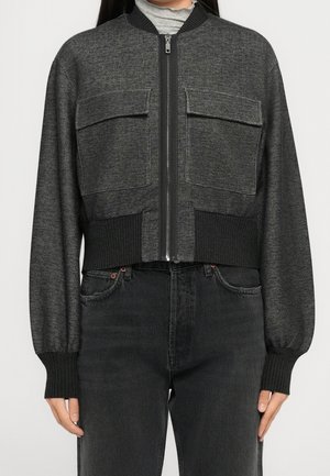 Kvinde iført en mørkegrå zip-up bomberjakke med store forlommer og sorte ribstrikkede manchetter og linning over sorte jeans.