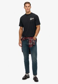 Camiseta de algodón negra con logo de texto blanco, combinada con jeans de mezclilla azul oscuro. Una camisa de cuadros roja está atada en la cintura. Se llevan mocasines marrones.