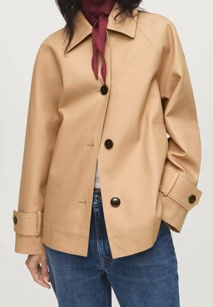 Kvinde iført en beige jakke med knapper og manchetstropper, blå jeans og et vinrødt tørklæde bundet om halsen mod en ensfarvet baggrund.