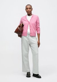 Femeie în picioare, îmbrăcată cu un cardigan roz, o cămașă albă, pantaloni largi gri deschis și pantofi negri, ținând o geantă mare maro sub braț.