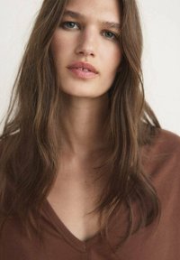 Smeđi v-izrez majica s mekom teksturom. Model ima dužu, valovitu kosu, neutralan make-up i neutralnu pozadinu, fokusirajući se na karakteristike lica.