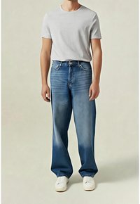 Homme portant des baskets blanches, un jean bleu décontracté et un t-shirt uni gris clair à manches courtes, debout devant un fond neutre.