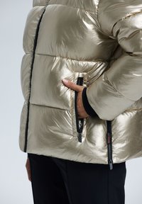 Metallisk guld puffjacka med en glänsande yta, dragkedjefickor och svarta dragkedjor, med en kviltad design.