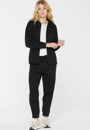 Blonde woman wearing black zip-up hoodie, black jogger pants, white shirt, and white sneakers, standing against a plain white background.