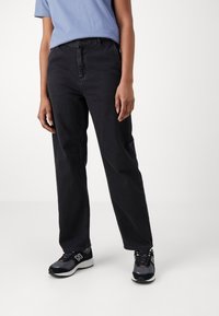 Zwarte denim broek met een rechte snit, voorzien van een knoop- en ritsluiting aan de voorkant, gecombineerd met zwart-grijze sportschoenen.
