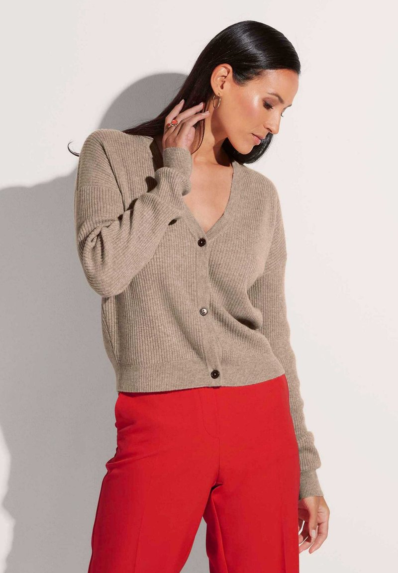Beige geribbelde cardigan met een diepe V-hals, lange mouwen en knopen aan de voorkant, gecombineerd met levendige rode broek.