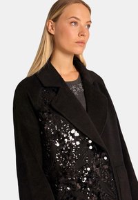 Cappotto nero realizzato in un tessuto morbido e testurizzato, con un design di paillettes su un lato. I dettagli notevoli includono un ampio colletto e tasche profonde.