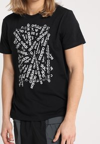 Musta puuvillainen t-paita, jossa on rohkea graafinen suunnittelu, jossa toistuu valkoinen "adidas" -teksti eri suuntiin ja suorakulmainen ääriviiva.